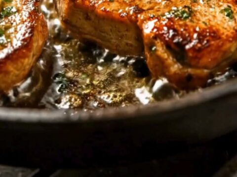 Four golden seared pork chops in a cast-iron skillet, topped with melting dollops of garlic butter and fresh parsley.