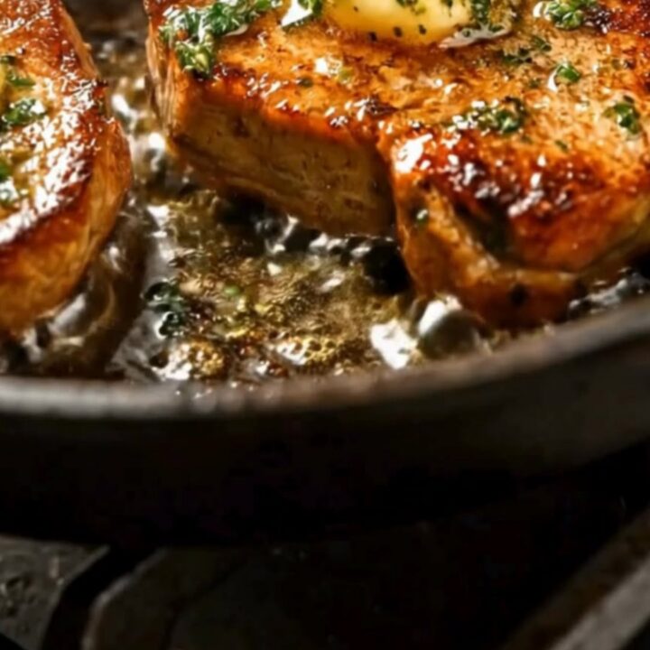 Four golden seared pork chops in a cast-iron skillet, topped with melting dollops of garlic butter and fresh parsley.