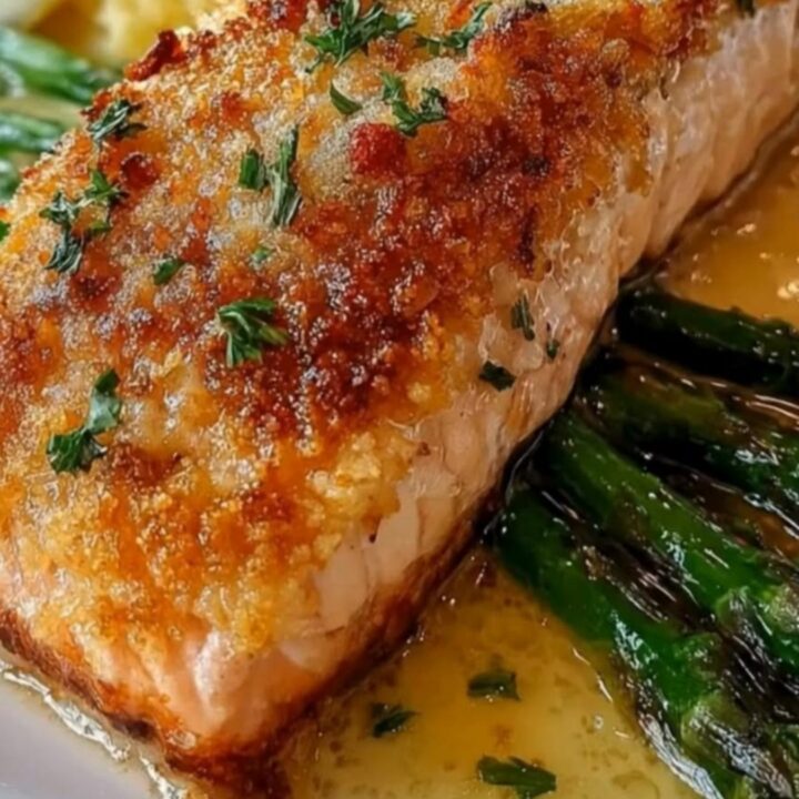 A golden-brown parmesan crusted salmon fillet served with glossy asparagus and lemon butter sauce on a white plate