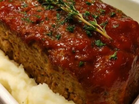 A whole glazed meatloaf garnished with fresh thyme, nestled in creamy mashed potatoes inside a white baker.