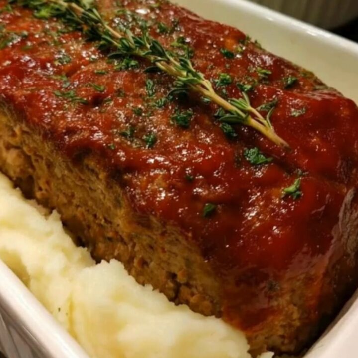 A whole glazed meatloaf garnished with fresh thyme, nestled in creamy mashed potatoes inside a white baker.