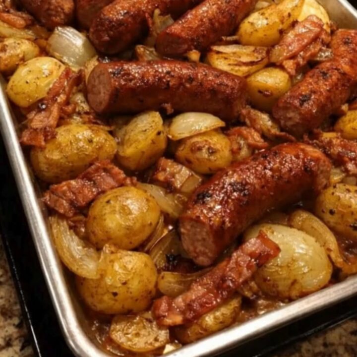 A sizzling sheet pan featuring browned sausage links, crispy bacon pieces, golden baby potatoes, and caramelized onions.