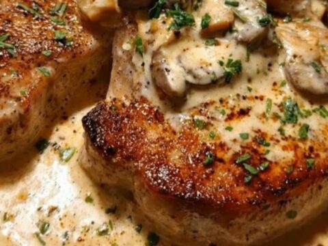 A seared pork chop smothered in a thick mushroom cream sauce and garnished with fresh parsley in a dark skillet