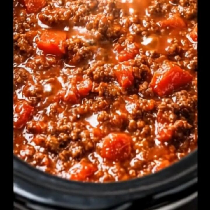 A black slow cooker filled to the brim with thick, glistening red meat sauce and tender tomato chunks.