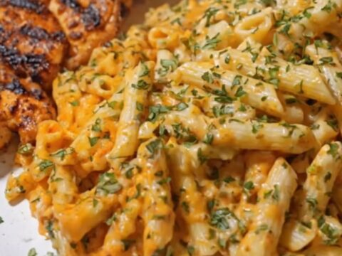 A white plate of creamy penne pasta with a massive cheese pull from a fork, topped with sliced charred Cajun chicken.