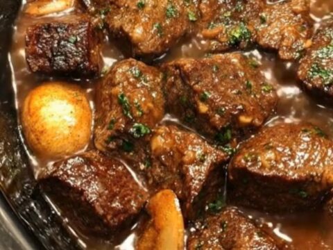 A black oval crock pot filled with glossy slow-cooked beef tips and tender mushrooms in rich brown gravy.
