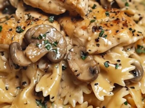 A white bowl filled with creamy farfalle pasta, seared chicken pieces, and sautéed mushrooms with steam rising.