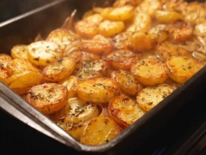 A dark roasting dish filled with golden-brown, herb-seasoned roasted potato rounds and caramelized onions.
