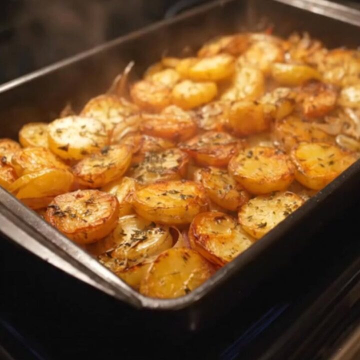 A dark roasting dish filled with golden-brown, herb-seasoned roasted potato rounds and caramelized onions.