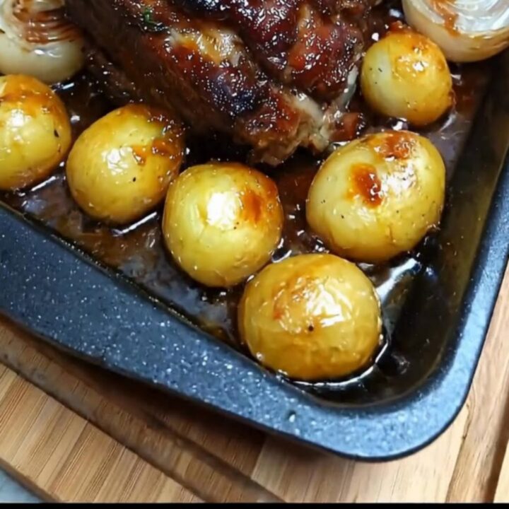 A succulent honey glazed roast pork topped with fresh rosemary in a roasting pan beside caramelized baby onions