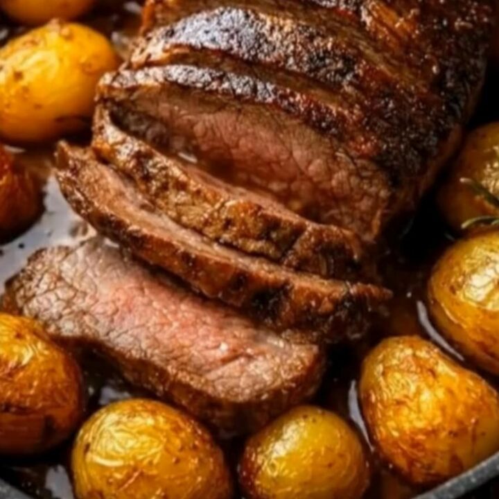 Slices of juicy garlic herb roast beef surrounded by crispy golden baby potatoes in a dark skillet.