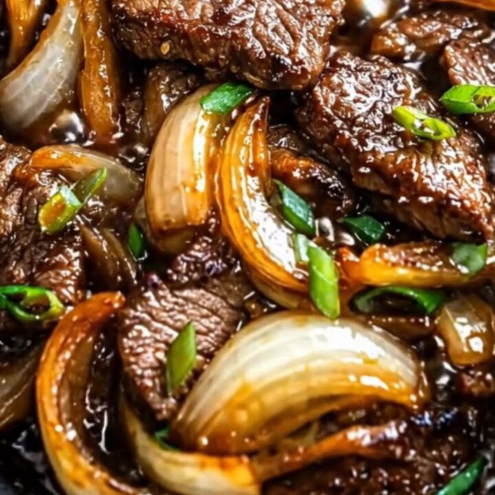 Glistening strips of tender beef and caramelized yellow onions tossed in a glossy brown sauce in a dark pan with steam