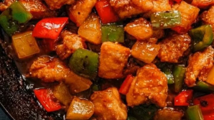 A rustic black skillet filled with glistening crispy pineapple chicken, red and green bell peppers