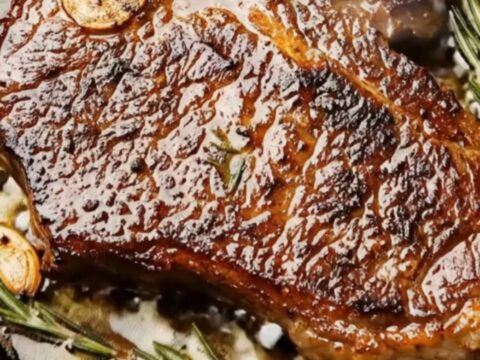 A glistening pan-seared T-bone steak resting in a cast-iron skillet with charred garlic and fresh rosemary sprigs.