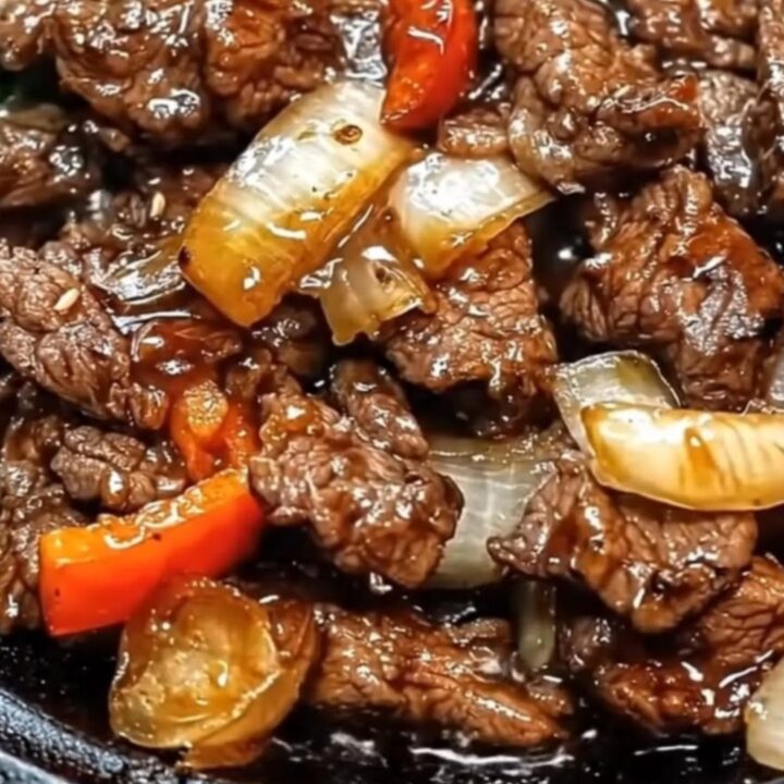 A hot cast iron platter filled with glistening strips of seared beef, sliced onions, and red peppers in a rich glaze.