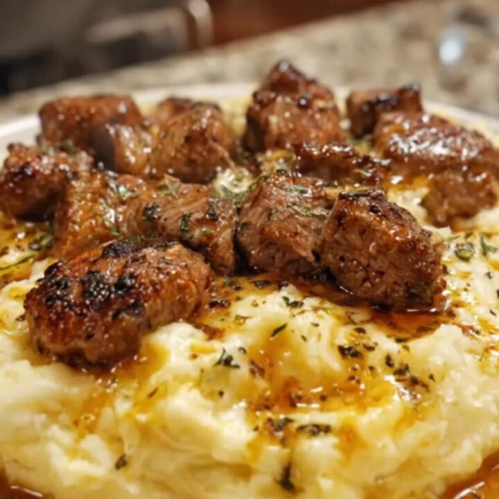 A white plate featuring tender, seared garlic butter steak bites piled on top of velvety mashed potatoes and drizzled