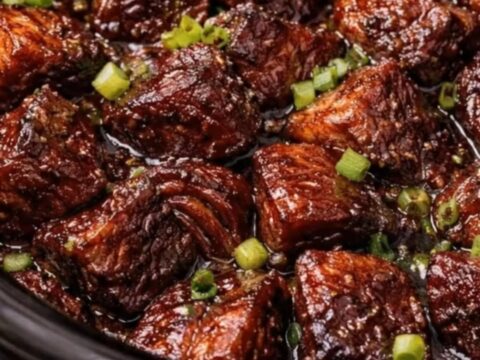 A close-up shot of caramelized, sticky BBQ beef burnt ends garnished with fresh green onions inside a black slow cooker.