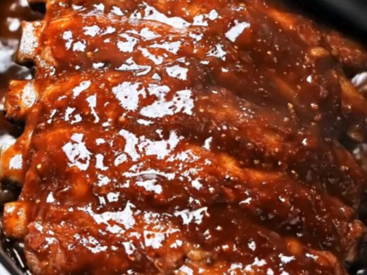 A close-up shot of glossy, sticky barbecue slow cooker ribs inside the black slow cooker bowl with steam rising.