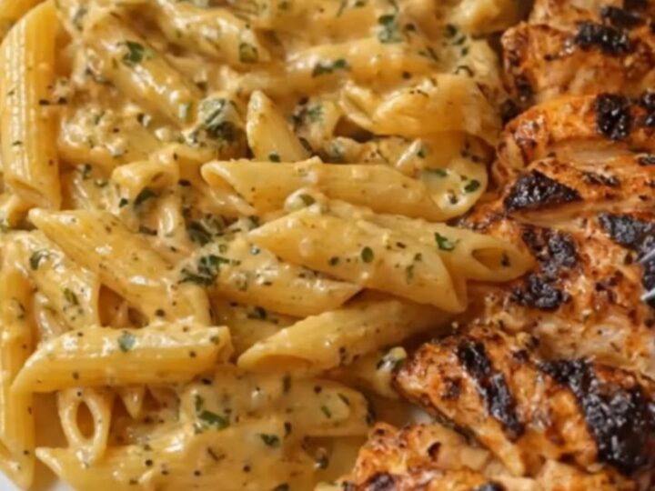 A white plate featuring creamy, glossy penne pasta alongside charred, sliced Cajun chicken breasts