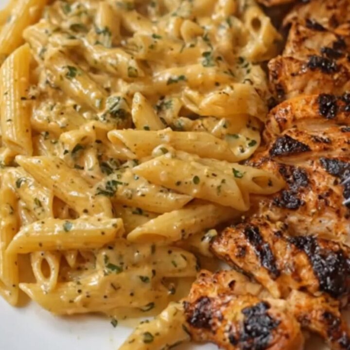 A white plate featuring creamy, glossy penne pasta alongside charred, sliced Cajun chicken breasts
