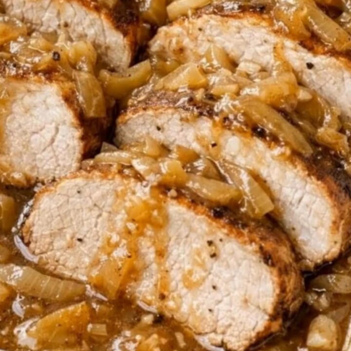 A white ceramic baking dish filled with juicy sliced pork tenderloin medallions smothered in glossy caramelized onion gravy.