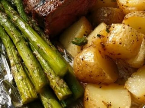 A wooden board holding an opened foil packet filled with juicy steak medallions, roasted potatoes, and asparagus.