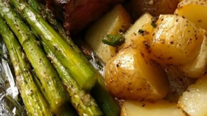 A wooden board holding an opened foil packet filled with juicy steak medallions, roasted potatoes, and asparagus.