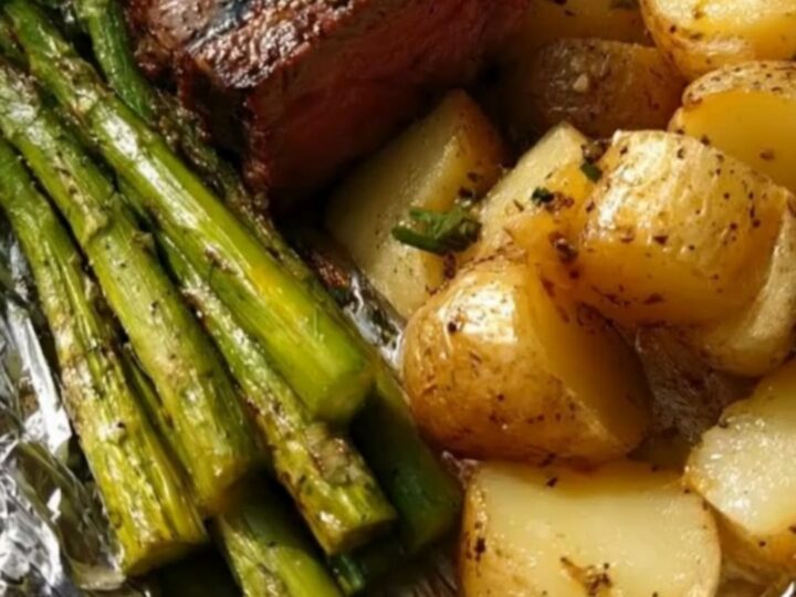 A wooden board holding an opened foil packet filled with juicy steak medallions, roasted potatoes, and asparagus.