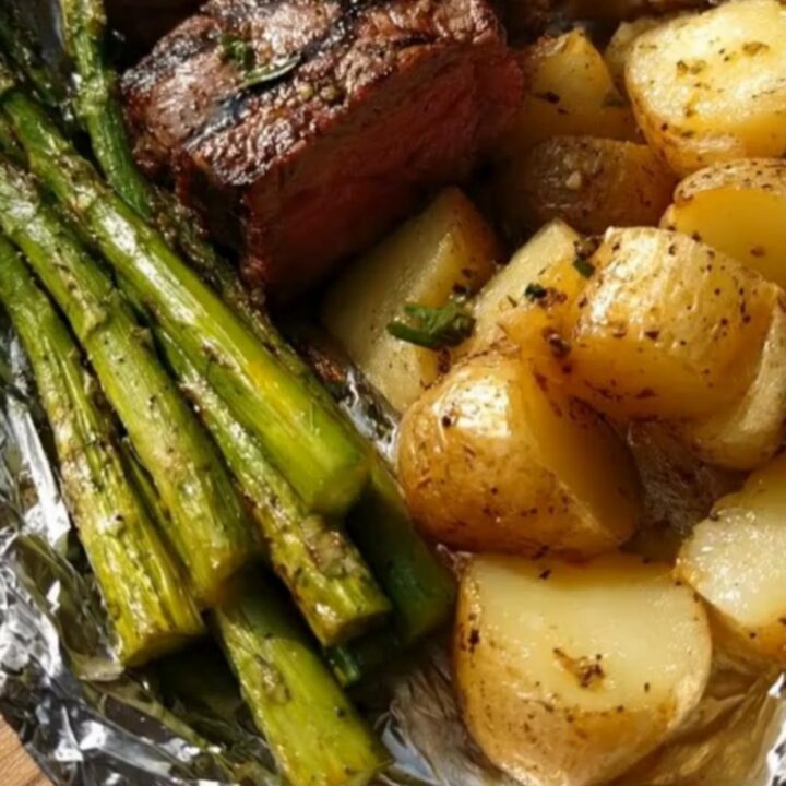 A wooden board holding an opened foil packet filled with juicy steak medallions, roasted potatoes, and asparagus.