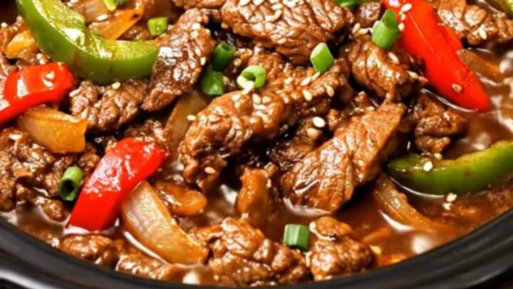 A close-up of tender beef strips and vibrant bell peppers in a glossy brown sauce inside a slow cooker.