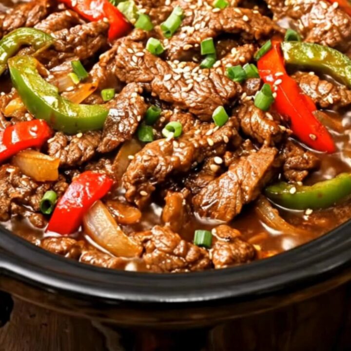 A close-up of tender beef strips and vibrant bell peppers in a glossy brown sauce inside a slow cooker.