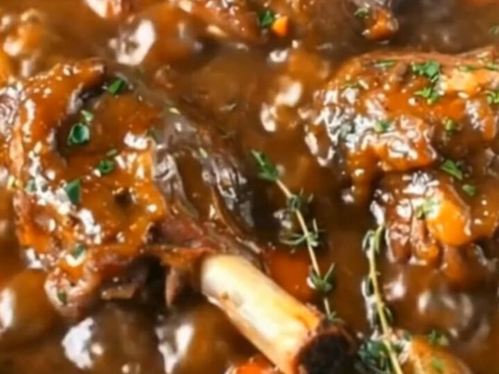 Slow-braised lamb shanks simmering in a rich, dark savory gravy with carrots, baby potatoes, and fresh thyme