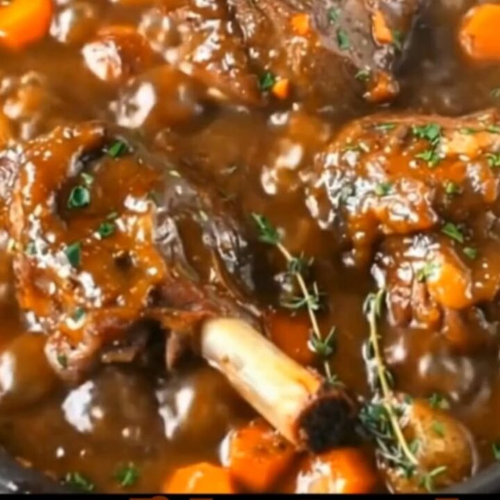 Slow-braised lamb shanks simmering in a rich, dark savory gravy with carrots, baby potatoes, and fresh thyme