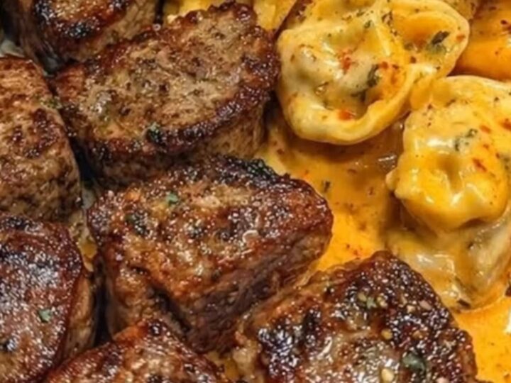 A plate of creamy, orange-hued tortellini Alfredo topped with glistening, seared garlic butter steak bites.
