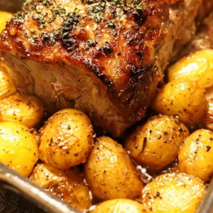 A roasted pork loin surrounded by glistening, golden-brown baby potatoes in a roasting pan