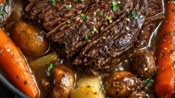 A dark Dutch oven containing sliced, tender beef pot roast surrounded by glistening glazed carrots and mushrooms