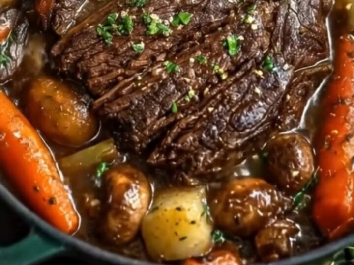 A dark Dutch oven containing sliced, tender beef pot roast surrounded by glistening glazed carrots and mushrooms