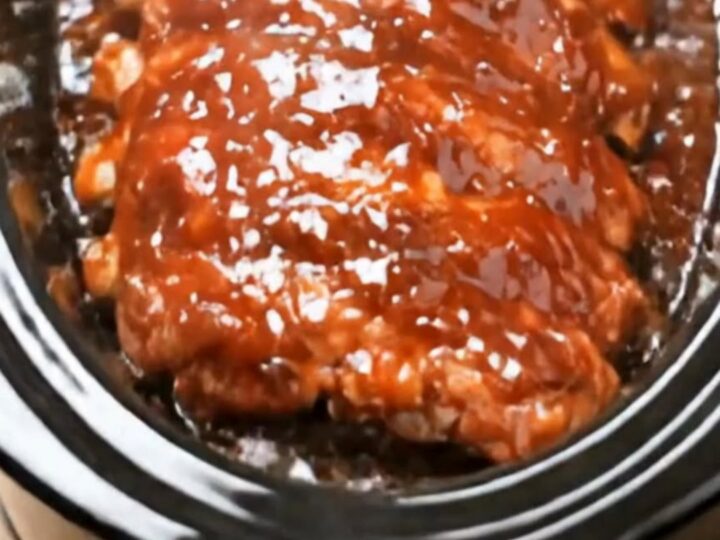 A rack of glossy, saucy crockpot BBQ ribs resting in a black oval slow cooker, looking extremely tender and delicious.