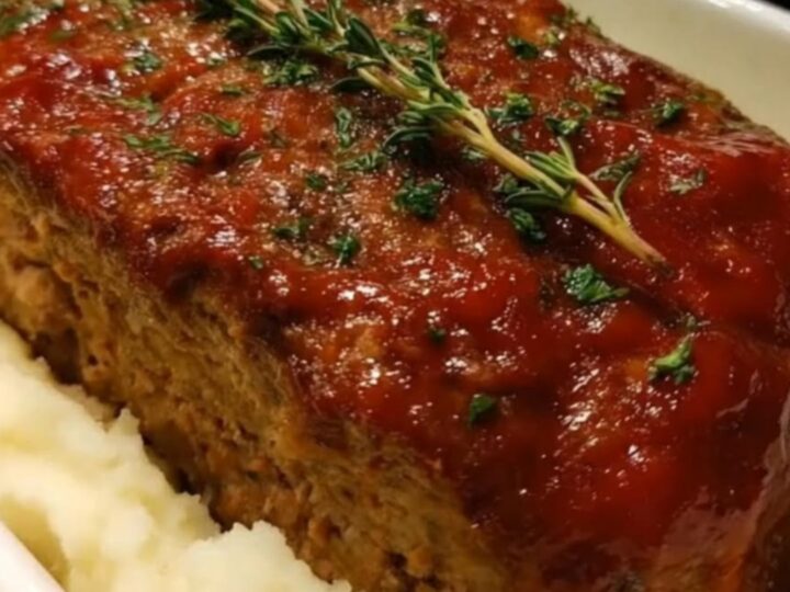 A glistening glazed meatloaf resting in a white baking dish on a bed of fluffy mashed potatoes, garnished with fresh thyme.