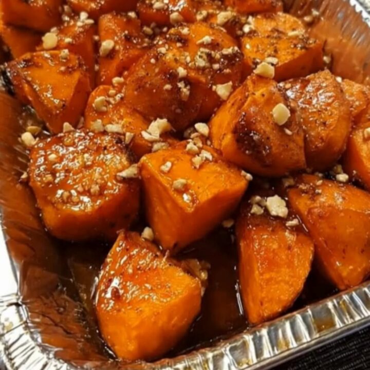 A warm foil pan filled with glossy, steaming sweet potatoes coated in bubbling brown sugar glaze and toasted pecans.
