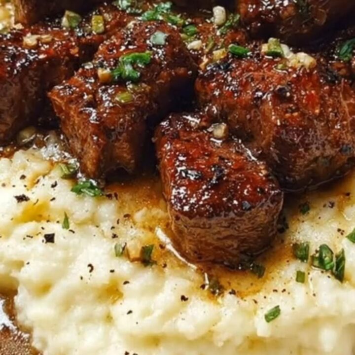Tender garlic butter steak bites piled high over a bed of creamy mashed potatoes in a white shallow bowl