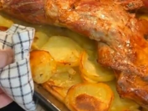 Slow-roasted lamb shoulder resting over golden sliced potatoes in a rustic roasting pan under warm kitchen lighting