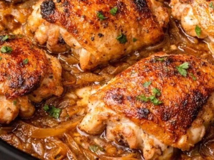 Six golden-brown chicken thighs resting on glossy caramelized onion gravy inside a black slow cooker garnished with fresh