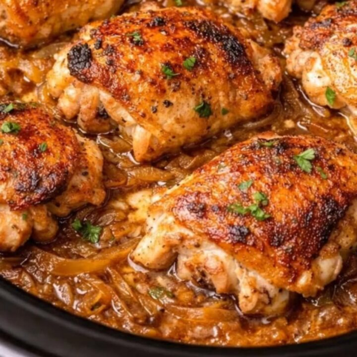 Six golden-brown chicken thighs resting on glossy caramelized onion gravy inside a black slow cooker garnished with fresh