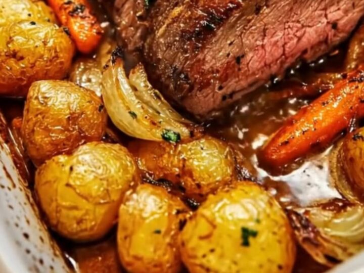 A beautiful white ceramic baking dish containing sliced garlic herb roast beef surrounded by tender baby potatoes