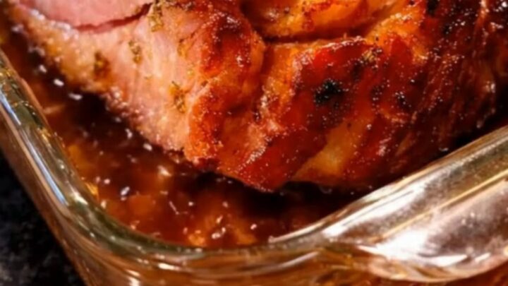 A beautifully glazed baked ham with a glistening caramelized crust, resting in a glass baking dish with rich pan juices.