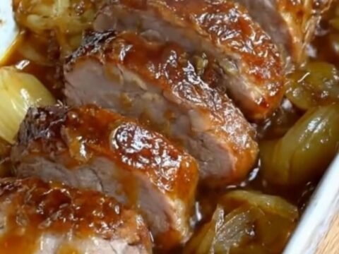 Sliced tender pork tenderloin medallions in a white baking dish surrounded by rich caramelized onions and a glossy jus