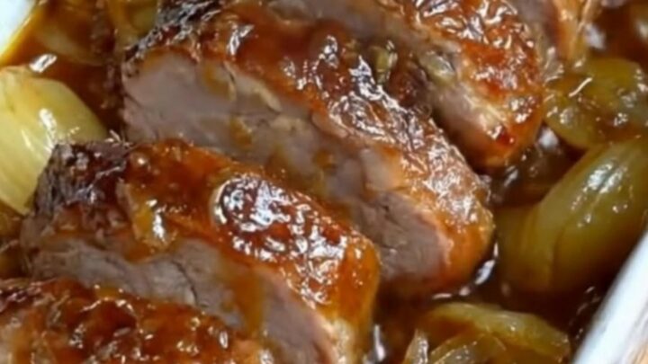 Sliced tender pork tenderloin medallions in a white baking dish surrounded by rich caramelized onions and a glossy jus