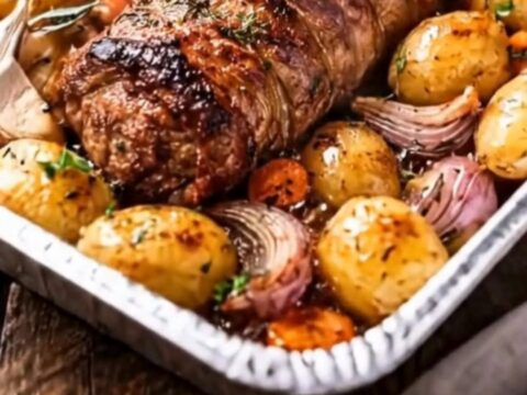 A golden-brown garlic herb pork loin roast sliced on a wooden board surrounded by roasted potatoes and onions.