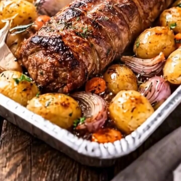 A golden-brown garlic herb pork loin roast sliced on a wooden board surrounded by roasted potatoes and onions.